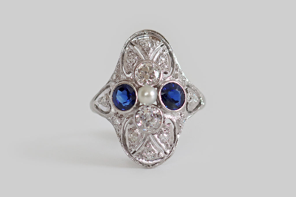 Antique Jewelry Portland, Vintage Jewelry Portland , Antique Engagement Rings | Poor Mouchette | A charming, Art-Deco-era, filigree shield ring, modeled in platinum, whose primary gems are arranged to suggest a quatrefoil. These gems— two natural, vibrant-blue sapphires, and two old European cut diamonds— are held in bezels at the center of the ring face, where they surround a single, natural seed pearl. This quatrefoil rests amid the ornate, hand-pierced, diamond-set forms that decorate the body 