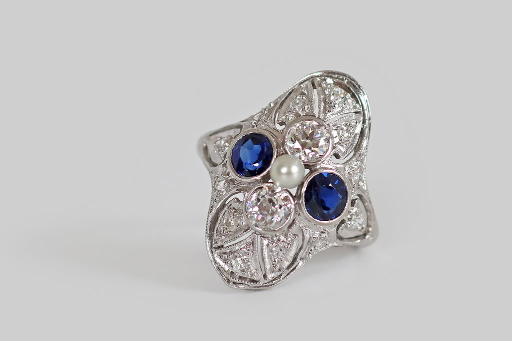 Antique Jewelry Portland, Vintage Jewelry Portland , Antique Engagement Rings | Poor Mouchette | A charming, Art-Deco-era, filigree shield ring, modeled in platinum, whose primary gems are arranged to suggest a quatrefoil. These gems— two natural, vibrant-blue sapphires, and two old European cut diamonds— are held in bezels at the center of the ring face, where they surround a single, natural seed pearl. This quatrefoil rests amid the ornate, hand-pierced, diamond-set forms that decorate the body 