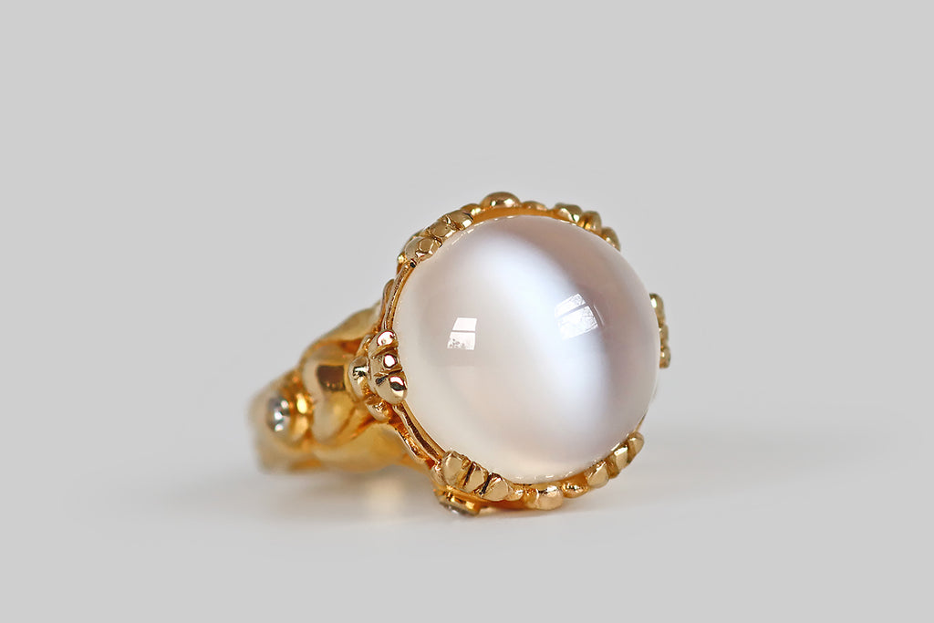 Poor Mouchette | Curated Antique Jewelry, Vintage Jewelry & Engagement Rings | A big, soulful, 1970s cocktail ring, modeled in 18k yellow gold, that features a magical, round cat's eye moonstone cabochon! This glowing white moonstone is held in six clusters of triple-prongs, where it reads like a crystal ball, sitting atop a spiritualist's stand. The moonstone's two, striking, adularescent bands have a subtle blue flash, and these "eyes" shift across its high dome, with movement.