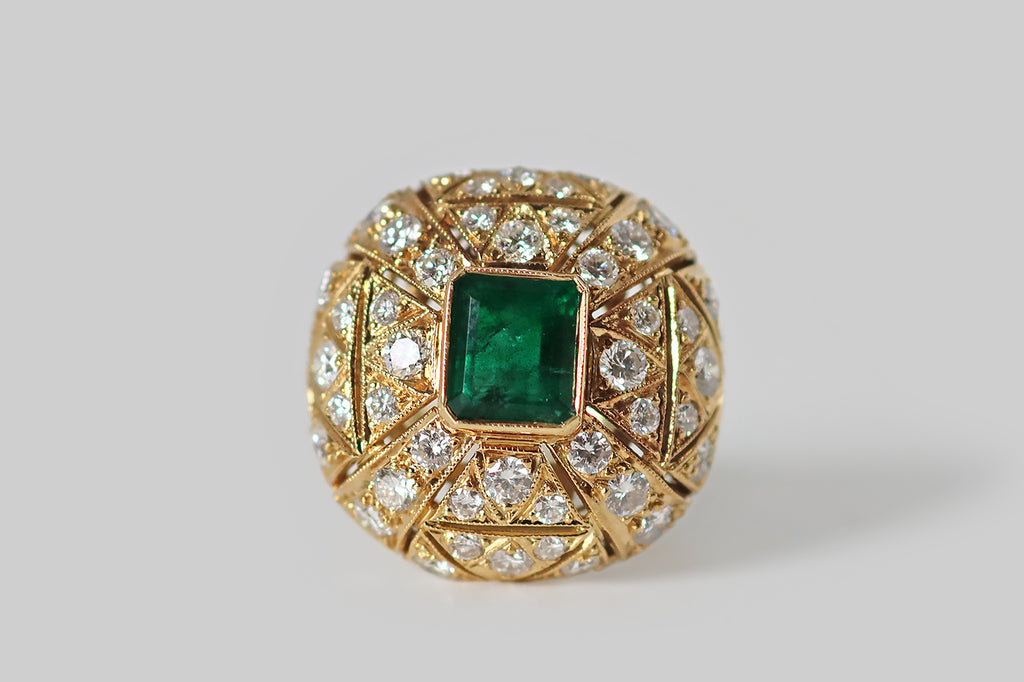 Antique Jewelry Portland, Vintage Jewelry Portland , Antique Engagement Rings | Poor Mouchette | A glamorous, mid-twentieth-century ring, modeled in 18k yellow gold, whose cushion-shaped face is encrusted with an ornate arrangement of gemstones— many, sparkling round brilliant cut diamonds, surrounding a central, rich-green, step-cut emerald.&nbsp;This ring has a soft, bombe-like profile. The emerald rests at the center of the ring face