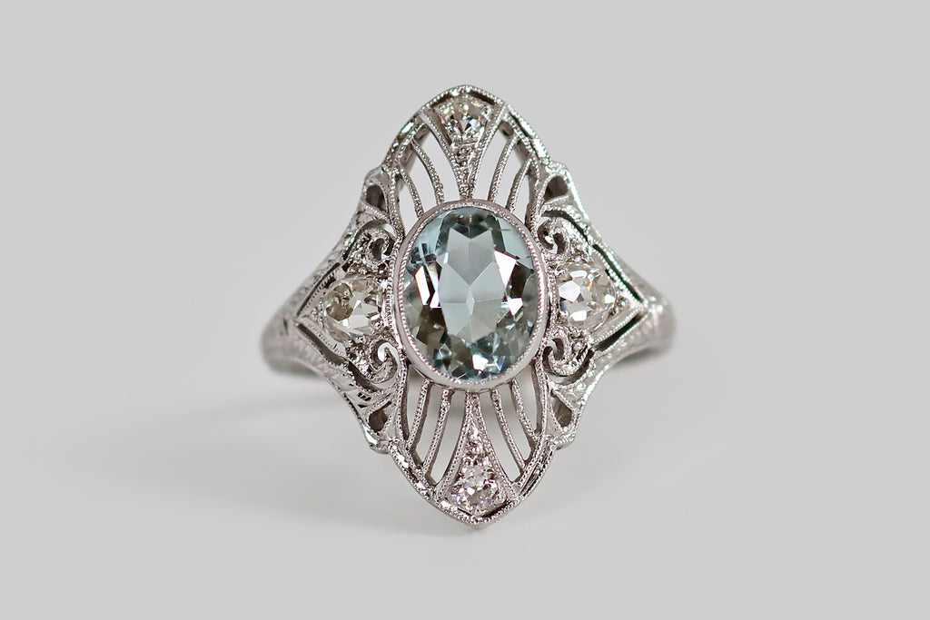 Antique Jewelry Portland, Vintage Jewelry Portland , Antique Engagement Rings | Poor Mouchette | An Art Deco era filigree shield ring, modeled in platinum, whose primary gem is a faceted, oval aquamarine. This icy aqua is held in a low-profile bezel, at the center of the ring face, where it is flanked by a pair of early old mine cut diamonds. Surrounding this trio of gems, an intricate, finely-executed, openwork design of knife-wire, curls, and paisleys. 