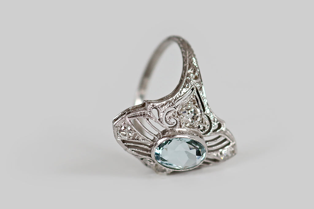 Antique Jewelry Portland, Vintage Jewelry Portland , Antique Engagement Rings | Poor Mouchette | An Art Deco era filigree shield ring, modeled in platinum, whose primary gem is a faceted, oval aquamarine. This icy aqua is held in a low-profile bezel, at the center of the ring face, where it is flanked by a pair of early old mine cut diamonds. Surrounding this trio of gems, an intricate, finely-executed, openwork design of knife-wire, curls, and paisleys. 