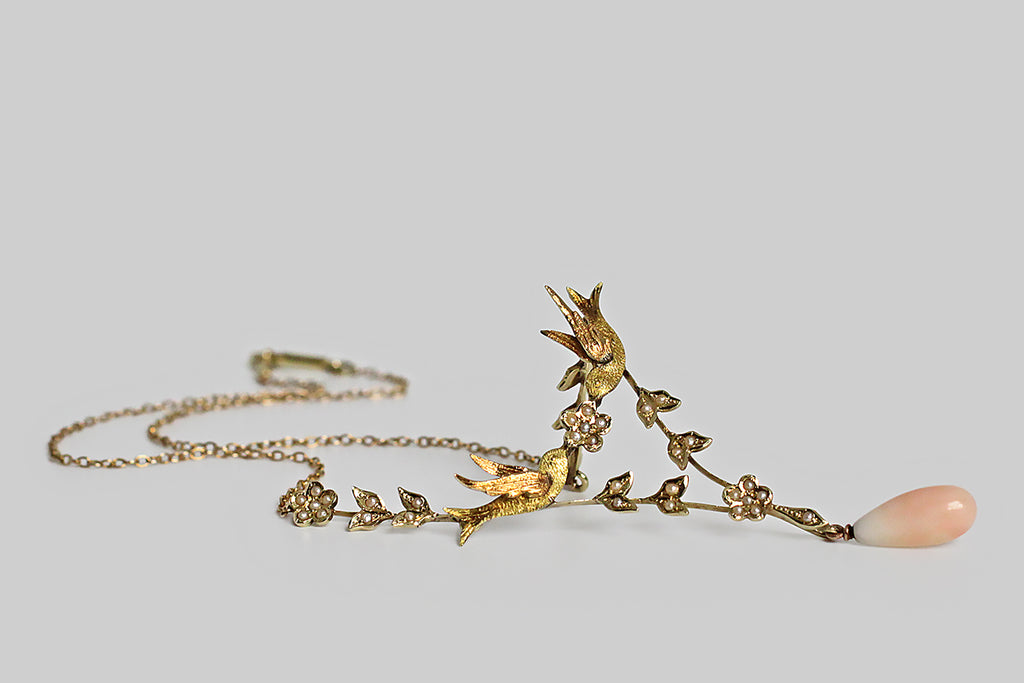 A Victorian-era necklace, with a sweet, serene presence. At its center, a pair of three-dimensional swallows come together to hold a five-petaled, pearl-set flower. An elegant, v-shaped framework of flowers and leaves is built beneath these bird friends, and a pair of jointed flowers attach them, from above, to the necklace's fine cable chain. All of the floral elements are set with natural seed pearls.